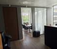 Office space with wooden flooring, large windows with blinds, a desk with chairs, a printer, and a reception area with a screen.