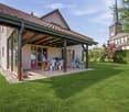 A well-maintained garden with a covered patio area, outdoor furniture, and a barbecue. The house has a modern design with large windows and a tiled roof.