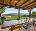 A wooden outdoor dining area with a table, chairs, and a grill, overlooking a scenic valley with mountains in the background.