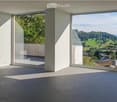 Bright, minimalist room with expansive windows offering panoramic views of a green landscape and distant mountains. The space is unfurnished, featuring a smooth concrete floor and white walls.