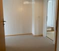 Empty hallway with carpeted floor, white paneled walls, and a ceiling light fixture. A doorway leads to a staircase area with wooden steps and a tiled landing.
