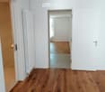 Empty hallway with wooden flooring, white walls, and a radiator on one side. A doorway leads to another room with similar flooring.