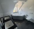 Modern bathroom with a corner bathtub, double sink vanity, and toilet. The space features dark tiled floors and white tiled walls, with a skylight window providing natural light.
