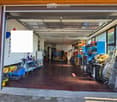 Open garage with wooden floor, various tools and equipment, large windows, and a partially open garage door leading to a paved outdoor area.