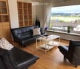 Modern living room with large windows offering a scenic view, featuring a black leather sofa, armchair, white coffee table, and wooden shelving unit.