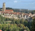 A panoramic view of a historic town with red-roofed buildings, a prominent church tower, and a modern suspension bridge spanning a river valley.