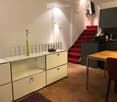 A modern, well-lit room with a wooden table, a chair, a white cabinet with glass doors, a green vase on the cabinet, and a staircase with red carpeting leading upwards.