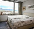 A well-lit bedroom with a large window offering a scenic view, featuring a neatly made bed with a floral duvet, a nightstand, a small plant on the windowsill, and decorative picture frames on the wall.
