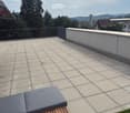 Rooftop terrace with tiled floor, low concrete wall, metal railing, and a wooden bench with a grey cushion.