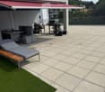 A modern outdoor patio with a sun lounger, a small seating area, a grill, and a storage box, featuring a tiled floor and a red awning.