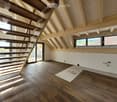 Empty room with wooden flooring, exposed wooden beams, and a staircase leading upwards. Large windows provide natural light, and the space is unfurnished.