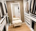 A modern walk-in closet with built-in white wardrobes, wooden flooring, a large mirror, a white fur-covered stool, and a decorative vase with dried branches.