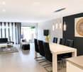 Modern open-plan living and dining area with a white dining table, black chairs, a black sectional sofa, a coffee table, and a large artwork on the wall. The space features a mix of white brick and dark walls, large windows with blinds, and modern pendant