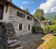 A rustic stone house with a sloped roof, wooden shutters, and a small garden area with vines and a gravel pathway.