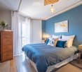 Cozy bedroom with blue walls, wooden floor, large bed with blue and white bedding, nightstands with lamps, dresser, mirror, and decorative vase on the dresser.