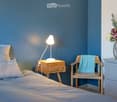 Cozy bedroom with blue walls, a neatly made bed, a wooden nightstand with a lamp, a small wooden chair with a blue towel, and a decorative vase with flowers on a dresser.
