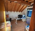 A furnished room with wooden beams on the ceiling, a desk with a laptop and screen, a chair, a small table with a cup, and a shelf with various items.