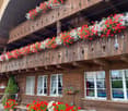 a wooden building with a balcony with flowers, a front bench, a desk, potted plants, white windows, and red flowers in front
