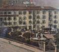 Hotel building with many windows, brown roof, garden in front, fountain, fence