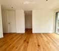 Empty room with light wooden flooring, white walls, and large windows offering a view of the outdoors.