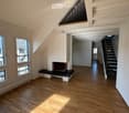 Bright, empty room with wooden flooring, large windows, and a fireplace. The space features a staircase leading to an upper level and a balcony area visible in the background.