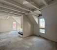 Empty attic space with exposed wooden beams, concrete floor, and large windows allowing natural light. Minimal furnishings include a small black bench and a ladder leading to another area.