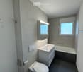 Modern bathroom with a toilet, sink, bathtub, and mirror. The room features grey tiled walls and a window providing natural light.