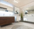 Modern bathroom with two sinks, one with a wooden cabinet and the other with white drawers, large mirrors with built-in lighting, and a decorative plant in a pot.