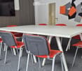 A modern, minimalist meeting or break room with a white rectangular table surrounded by colorful chairs, a large flat-screen TV mounted on the wall, and a decorative geometric wall design.