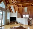 Bright living room with wooden beams and slanted ceiling, featuring a television on a wooden stand, a small wooden chair, a decorative sleigh, and a fur rug on the floor.