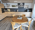 Modern kitchen with wooden cabinets, a round wooden dining table surrounded by wooden chairs, a pendant light fixture, and a sink area with a faucet. The space features a sleek design with a mix of light and dark tones.