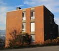 Modern two-story brick building with large windows and a flat roof, surrounded by greenery and shrubs.