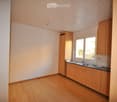 Bright, unfurnished kitchen with light wooden cabinets, a sink, and a countertop. The space features a wooden floor and large windows allowing natural light to enter.