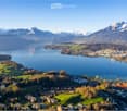 Aerial view of a scenic lakeside town surrounded by mountains, with a lake, green fields, and scattered buildings.