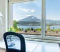 Bright room with large windows offering scenic mountain and lake views, furnished with a desk, chair, lamp, and flower vase.