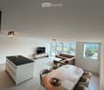 Modern living and dining area with a sofa, coffee table, dining table with chairs, and a kitchen island. Large windows offer a scenic view.