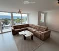 Modern living room with large windows offering a scenic view, featuring a sectional sofa, coffee table, and a small table with chairs on a balcony.