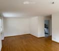 Empty room with wooden flooring, white walls, and a window on one side. A small kitchen area is visible through an open doorway.