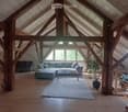 Spacious living room with exposed wooden beams, large windows, grey sectional sofa, coffee table, ottoman, and potted plants.