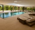 Modern indoor-outdoor lounge area with a swimming pool, large windows offering a lush green view, and comfortable seating including sofas, tables, and chairs.