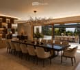 Elegant dining room with a long dark wooden dining table surrounded by beige upholstered chairs, a large chandelier, and decorative items on shelves. The room features a view of an outdoor dining area through large glass doors.