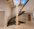 Spacious hallway with a modern glass staircase, a tall decorative light fixture, and a skylight above. The walls are light-colored, and the floor is tiled.