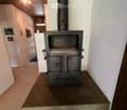 A wood-burning fireplace with a black chimney pipe, situated on a concrete platform in a room with white walls and a wooden ceiling.