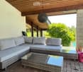 Outdoor patio with wooden ceiling, grey sectional sofa, glass coffee table, brick floor, and swimming pool visible in the background.