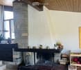 Cozy living room with a stone fireplace, wooden ceiling beams, and a large window. A small table and chair are positioned near the fireplace, along with decorative items like vases and a picture frame on the wall.