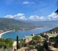 A scenic coastal view from a hillside property, featuring a house with a green roof, surrounded by lush greenery, overlooking a sandy beach and the Mediterranean Sea with distant mountains.
