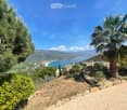 A scenic outdoor view from a hillside, featuring a brick pathway, lush greenery, a palm tree, and a distant coastal town with a beach and mountains in the background.