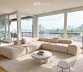 Modern living room with large windows offering a city view, featuring a beige sectional sofa, a marble coffee table, a floor lamp, and a dining area with chairs and a table.
