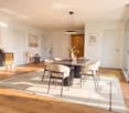 Modern dining room with a round wooden dining table, six chairs, a hanging pendant light, a side table with a lamp, and decorative vases on a cabinet.