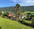 A picturesque rural landscape with a house, lush green fields, and a church in the distance, surrounded by trees and hills.
